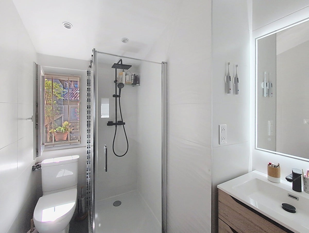 Salle d'eau entièrement carrelée en grand format blanc, équipée d'une douche à l'italienne avec paroi en verre, colonne de douche noire avec pommeau carré et douchette, et d'un meuble vasque suspendu en bois avec plan vasque blanc et miroir LED rectangulaire. Des WC suspendus sont installés contre le mur gauche sous une fenêtre ouvrant sur l'extérieur, avec un décor de mosaïque à reflets chromés en bande verticale.
