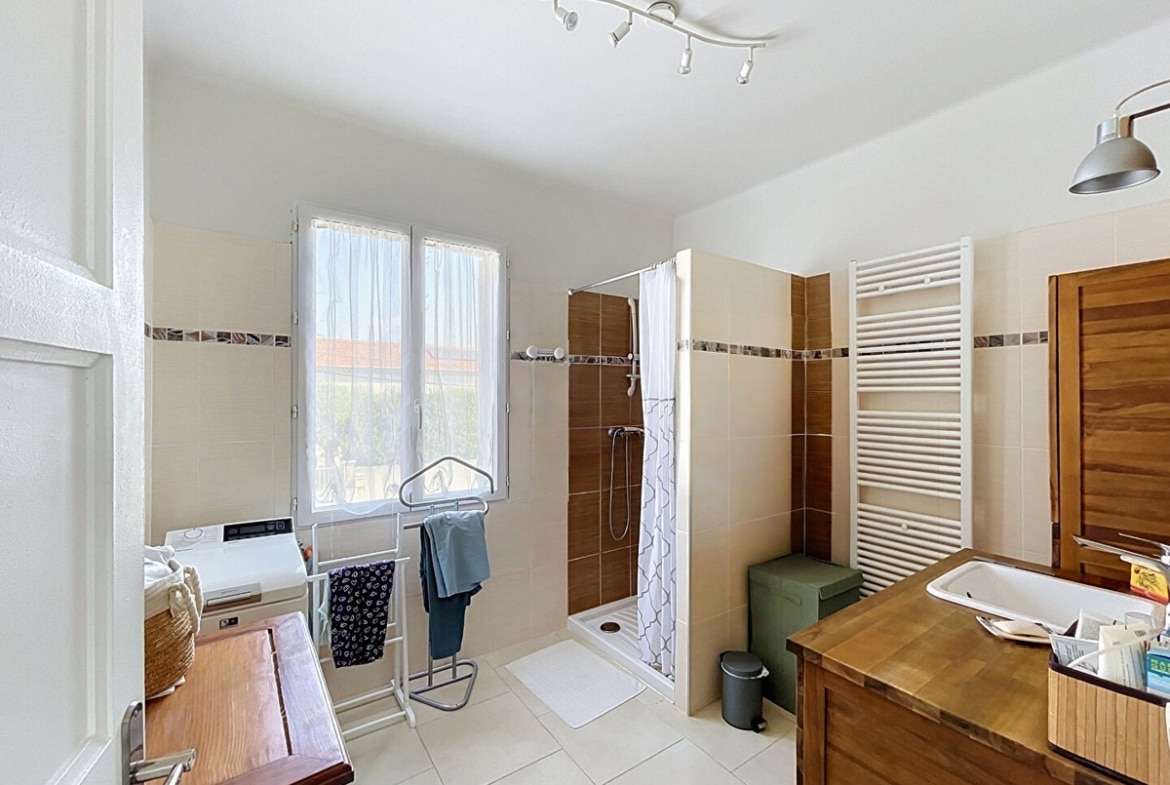 Salle de bain avec douche à l'italienne carrelée de grès brun et beige, meuble vasque en bois massif teck avec plan de travail, radiateur sèche-serviettes blanc et lave-linge intégré. Fenêtre PVC donnant sur l'extérieur, carrelage mural bicolore beige et brun avec frise décorative, sol en carrelage blanc.