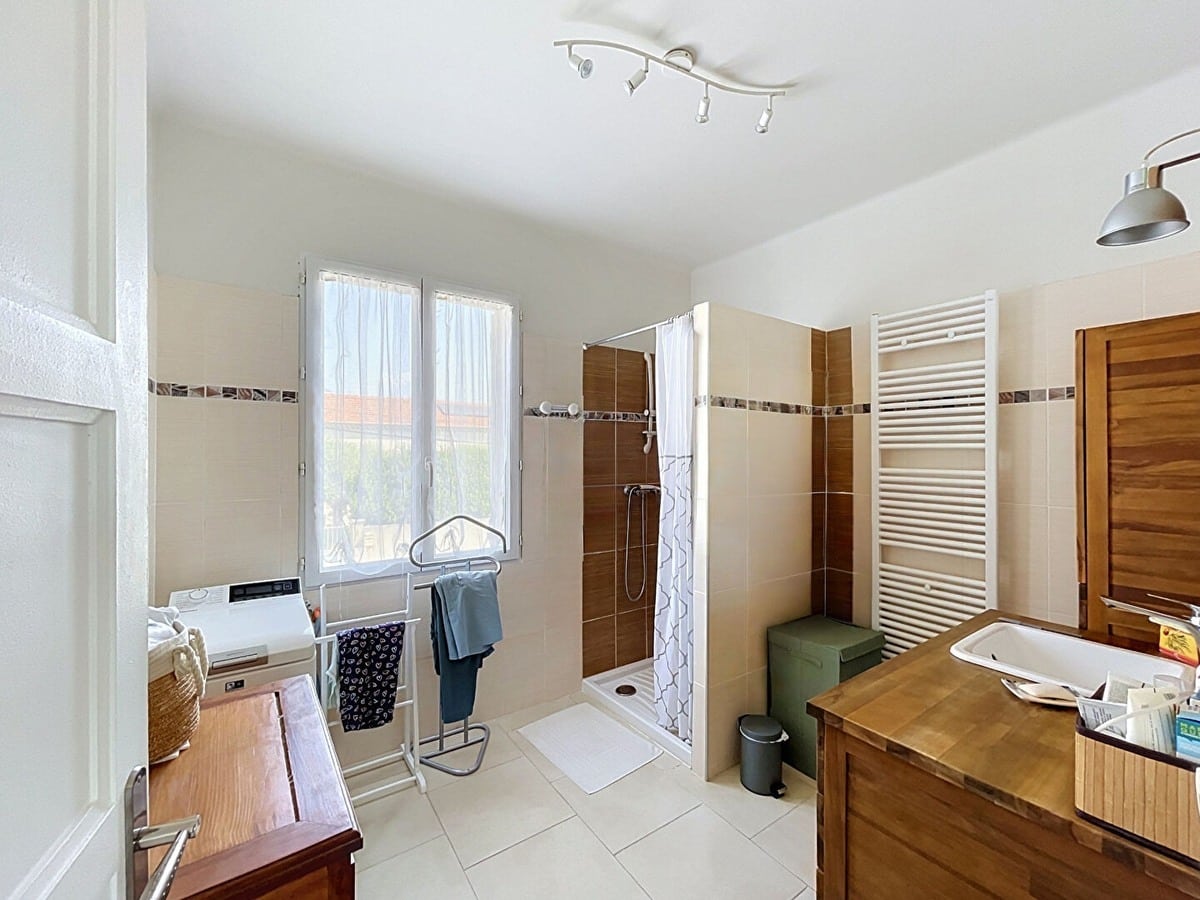 Salle de bain avec douche à l'italienne carrelée de grès brun et beige, meuble vasque en bois massif teck avec plan de travail, radiateur sèche-serviettes blanc et lave-linge intégré. Fenêtre PVC donnant sur l'extérieur, carrelage mural bicolore beige et brun avec frise décorative, sol en carrelage blanc.