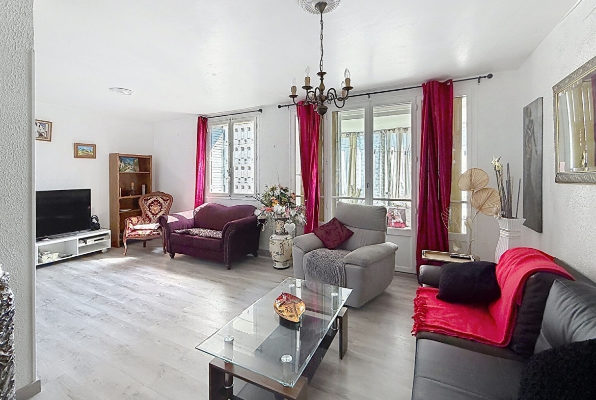 Séjour rectangulaire avec sol en parquet stratifié clair, équipé d'un canapé en cuir sombre, d'un canapé violet, d'un fauteuil Louis XV et d'une chaise grise, réunis autour d'une table basse en verre. Les fenêtres sont habillées de rideaux rouge fuchsia et un lustre en métal bronze à bougies est suspendu au plafond blanc.
