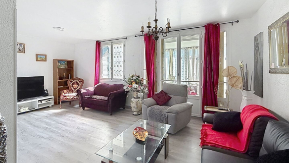 Séjour rectangulaire avec sol en parquet stratifié clair, équipé d'un canapé en cuir sombre, d'un canapé violet, d'un fauteuil Louis XV et d'une chaise grise, réunis autour d'une table basse en verre. Les fenêtres sont habillées de rideaux rouge fuchsia et un lustre en métal bronze à bougies est suspendu au plafond blanc.
