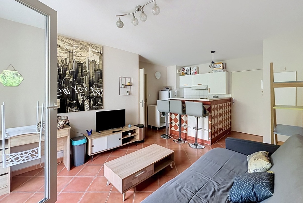 Pièce de vie avec sol en carreaux terre cuite, comprenant un canapé gris, une table basse en bois clair, un meuble TV bas bicolore et une télévision à écran plat, avec en fond un papier peint panoramique représentant la skyline de New York en noir et blanc. La cuisine ouverte est délimitée par un bar à carreaux en losanges rouge et blanc avec trois tabourets gris, et donne accès à la terrasse par une porte-fenêtre coulissante sur la gauche.