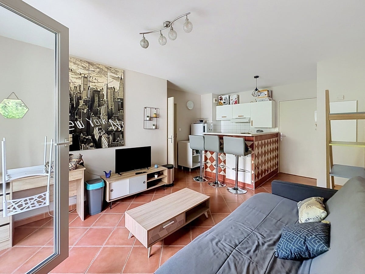 Pièce de vie avec sol en carreaux terre cuite, comprenant un canapé gris, une table basse en bois clair, un meuble TV bas bicolore et une télévision à écran plat, avec en fond un papier peint panoramique représentant la skyline de New York en noir et blanc. La cuisine ouverte est délimitée par un bar à carreaux en losanges rouge et blanc avec trois tabourets gris, et donne accès à la terrasse par une porte-fenêtre coulissante sur la gauche.