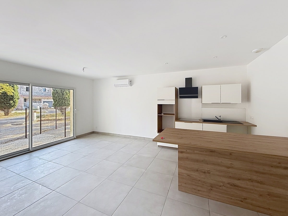 Pièce de vie ouverte avec carrelage gris clair grand format, cuisine intégrée aux façades blanches et plan de travail bois, équipée d'une hotte noire murale et d'un évier noir encastré. Un îlot central en bois est positionné au premier plan, une baie vitrée coulissante donne sur la cour gravillonnée, et un climatiseur mural blanc est installé en hauteur.
