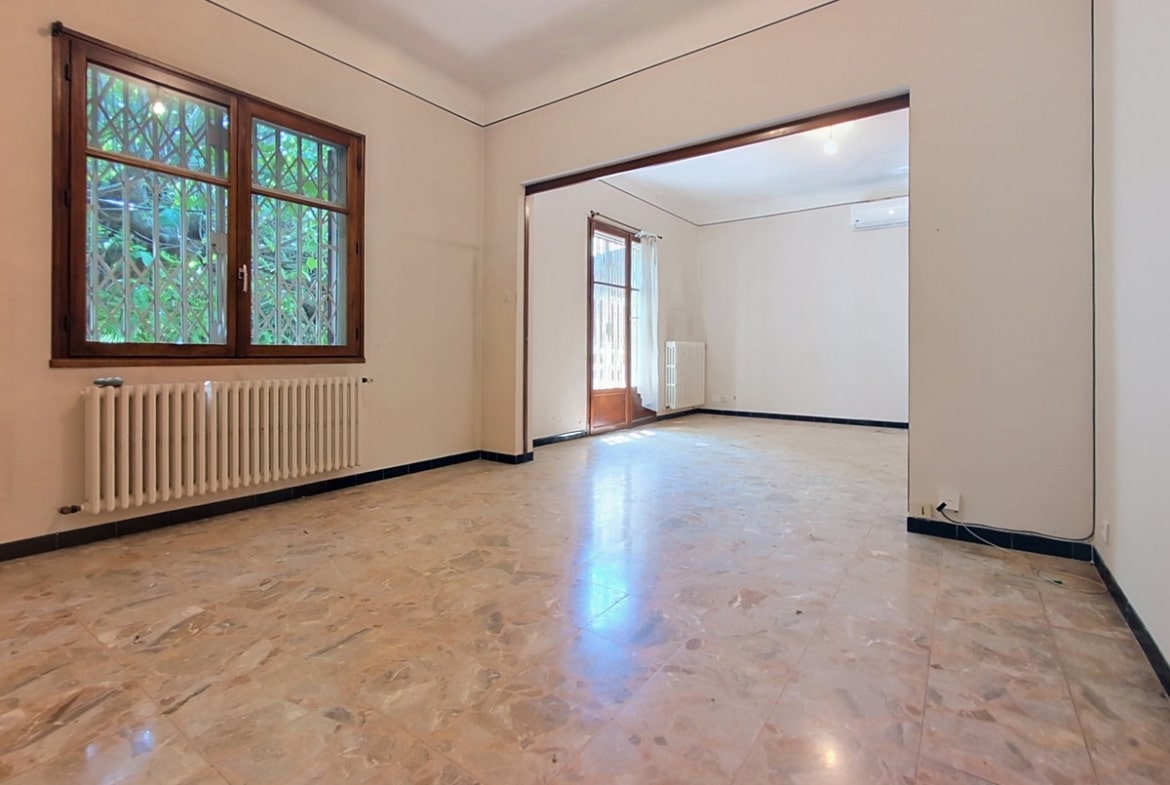 Double séjour communicant via une large ouverture en bois sombre, avec sol en dalles de marbre rose et beige poli, radiateurs en fonte blanche et fenêtres à croisillons avec volets repliables. La pièce de droite dispose d'un climatiseur mural blanc et d'une porte-fenêtre donnant sur l'extérieur.