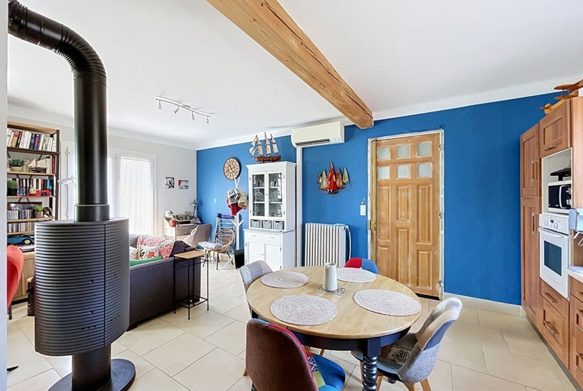 Séjour ouvert sur la cuisine avec un poêle à bois noir cylindrique au premier plan, mur peint en bleu cobalt orné de décorations marines dont une maquette de voilier et une horloge en forme de hublot. Canapé gris en fond, bibliothèque en bois chargée de livres, et buffet blanc vitré, sol en carrelage beige clair.