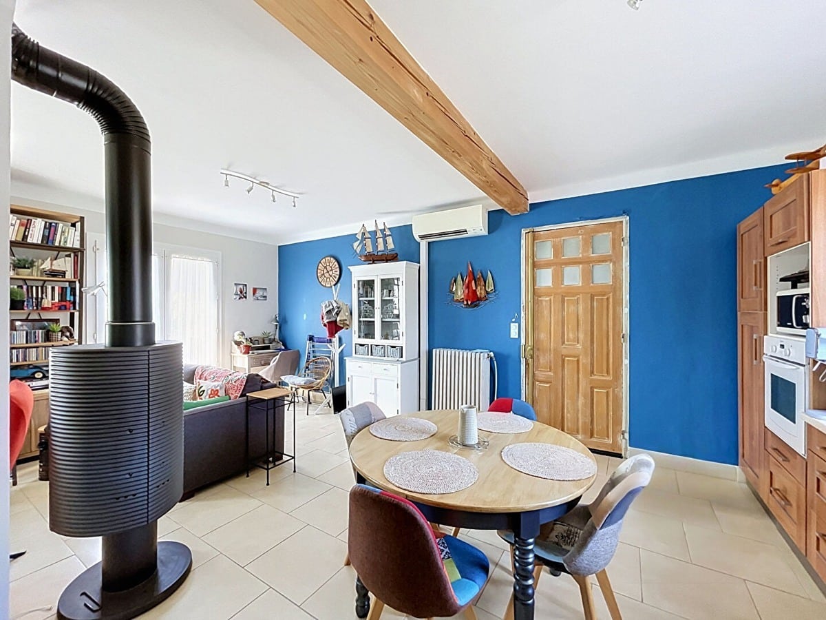 Séjour ouvert sur la cuisine avec un poêle à bois noir cylindrique au premier plan, mur peint en bleu cobalt orné de décorations marines dont une maquette de voilier et une horloge en forme de hublot. Canapé gris en fond, bibliothèque en bois chargée de livres, et buffet blanc vitré, sol en carrelage beige clair.