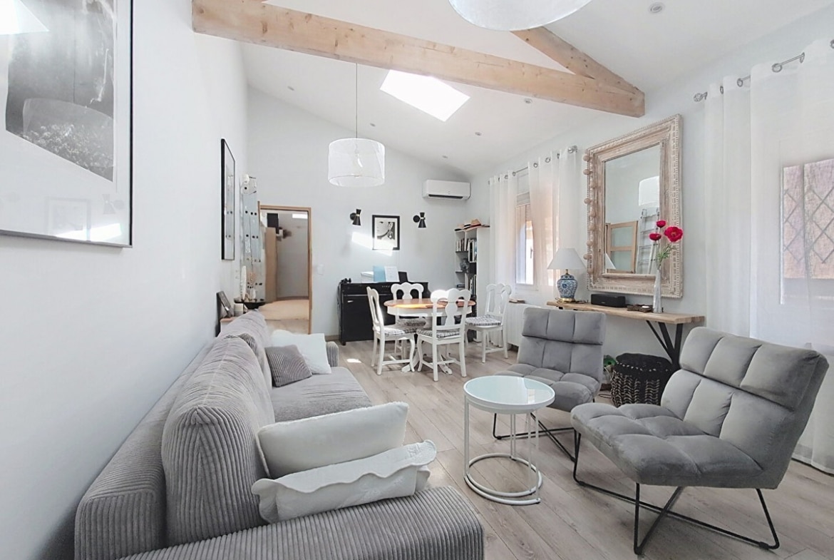Pièce de vie lumineuse aux murs blancs avec parquet stratifié clair, meublée d'un canapé gris en velours côtelé, d'un fauteuil gris et d'une table basse ronde en verre blanc, sous des poutres en bois naturel croisées en charpente apparente. Un espace salle à manger avec table ronde et chaises blanches est visible en arrière-plan, près d'un piano noir et d'un miroir doré à cadre sculpté.
