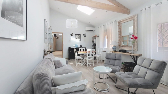 Pièce de vie lumineuse aux murs blancs avec parquet stratifié clair, meublée d'un canapé gris en velours côtelé, d'un fauteuil gris et d'une table basse ronde en verre blanc, sous des poutres en bois naturel croisées en charpente apparente. Un espace salle à manger avec table ronde et chaises blanches est visible en arrière-plan, près d'un piano noir et d'un miroir doré à cadre sculpté.