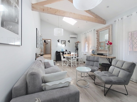 Pièce de vie lumineuse aux murs blancs avec parquet stratifié clair, meublée d'un canapé gris en velours côtelé, d'un fauteuil gris et d'une table basse ronde en verre blanc, sous des poutres en bois naturel croisées en charpente apparente. Un espace salle à manger avec table ronde et chaises blanches est visible en arrière-plan, près d'un piano noir et d'un miroir doré à cadre sculpté.