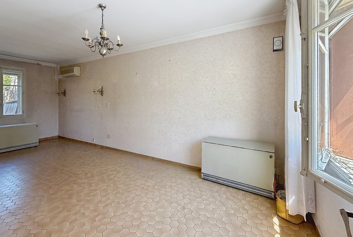 Pièce de séjour vide aux murs revêtus d'un papier peint beige texturé, avec un lustre chandelier en métal argenté à cinq bougies suspendu au plafond blanc. Le sol est recouvert de tomettes hexagonales couleur terre cuite, un radiateur à eau blanc de grande longueur est positionné le long du mur, une unité de climatisation réversible est fixée en hauteur et une porte-fenêtre à vitrage translucide éclaire le côté droit.