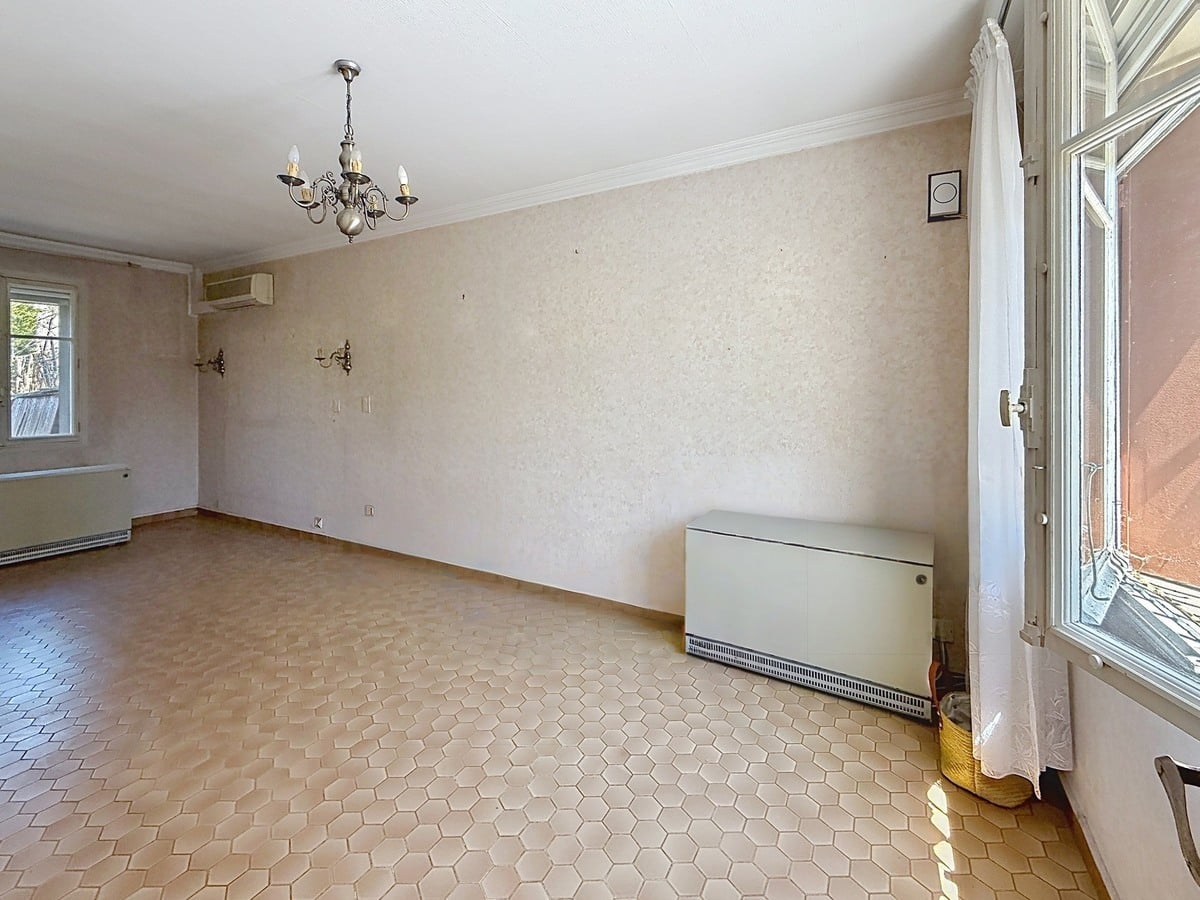 Pièce de séjour vide aux murs revêtus d'un papier peint beige texturé, avec un lustre chandelier en métal argenté à cinq bougies suspendu au plafond blanc. Le sol est recouvert de tomettes hexagonales couleur terre cuite, un radiateur à eau blanc de grande longueur est positionné le long du mur, une unité de climatisation réversible est fixée en hauteur et une porte-fenêtre à vitrage translucide éclaire le côté droit.