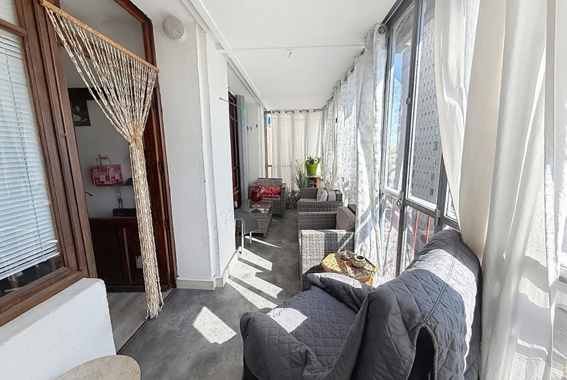 Loggia vitrée et couverte aménagée en salon d'extérieur avec canapé gris, fauteuils en résine tressée grise et table basse, desservie par une porte en bois avec rideau de perles. Les grandes baies vitrées donnent sur des bâtiments environnants et laissent entrer une lumière naturelle abondante.