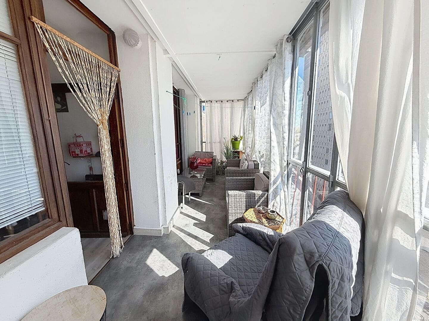 Loggia vitrée et couverte aménagée en salon d'extérieur avec canapé gris, fauteuils en résine tressée grise et table basse, desservie par une porte en bois avec rideau de perles. Les grandes baies vitrées donnent sur des bâtiments environnants et laissent entrer une lumière naturelle abondante.