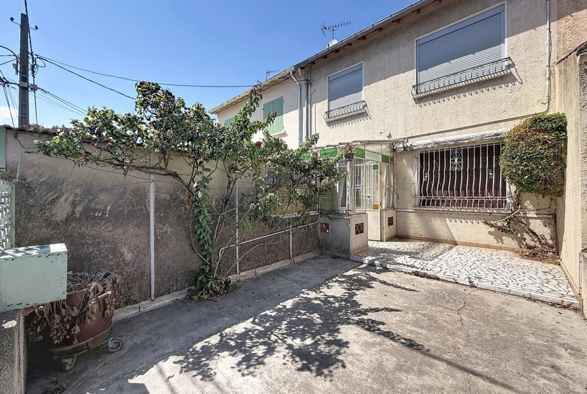 Terrasse avant en béton délimitée par un mur mitoyen, avec une végétation grimpante couvrant en partie le mur gauche et un arbre en espalier. L'entrée de la maison est accessible par quelques marches avec une rampe en fer forgé, une véranda ancienne à structure métallique vitrée marque le seuil d'entrée, et une grille de protection en fer forgé sécurise la fenêtre en rez-de-chaussée.