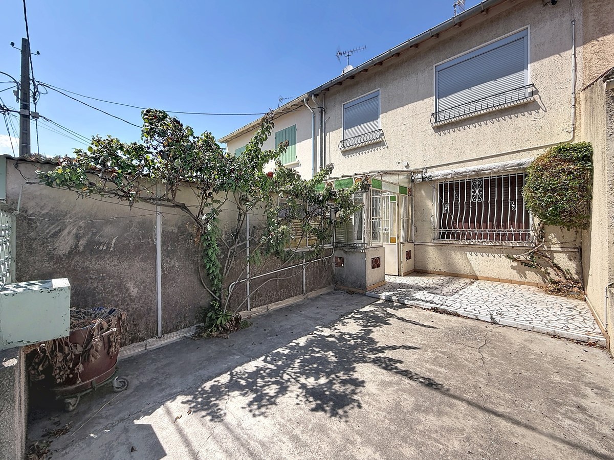 Terrasse avant en béton délimitée par un mur mitoyen, avec une végétation grimpante couvrant en partie le mur gauche et un arbre en espalier. L'entrée de la maison est accessible par quelques marches avec une rampe en fer forgé, une véranda ancienne à structure métallique vitrée marque le seuil d'entrée, et une grille de protection en fer forgé sécurise la fenêtre en rez-de-chaussée.