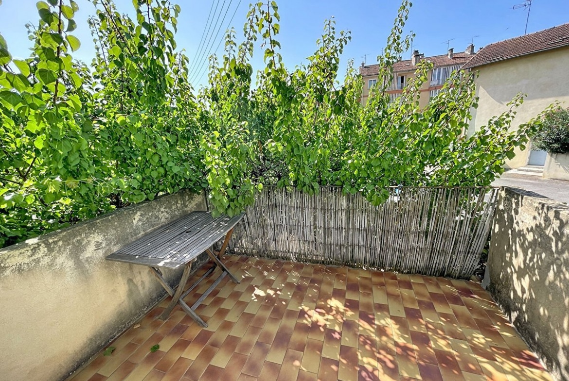 Terrasse en hauteur avec carrelage en damier ocre et rouge, entourée d'un muret en béton et d'un claustra en bambou recouvert de végétation grimpante touffue. Table de jardin en bois pliante installée dans un coin ombragé, ciel bleu dégagé visible au-dessus de la canopée.