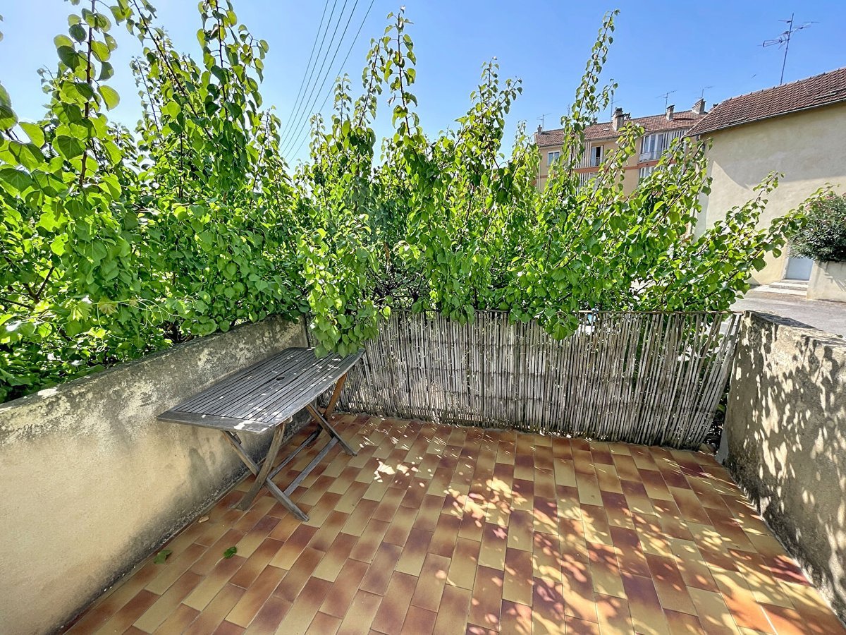 Terrasse en hauteur avec carrelage en damier ocre et rouge, entourée d'un muret en béton et d'un claustra en bambou recouvert de végétation grimpante touffue. Table de jardin en bois pliante installée dans un coin ombragé, ciel bleu dégagé visible au-dessus de la canopée.