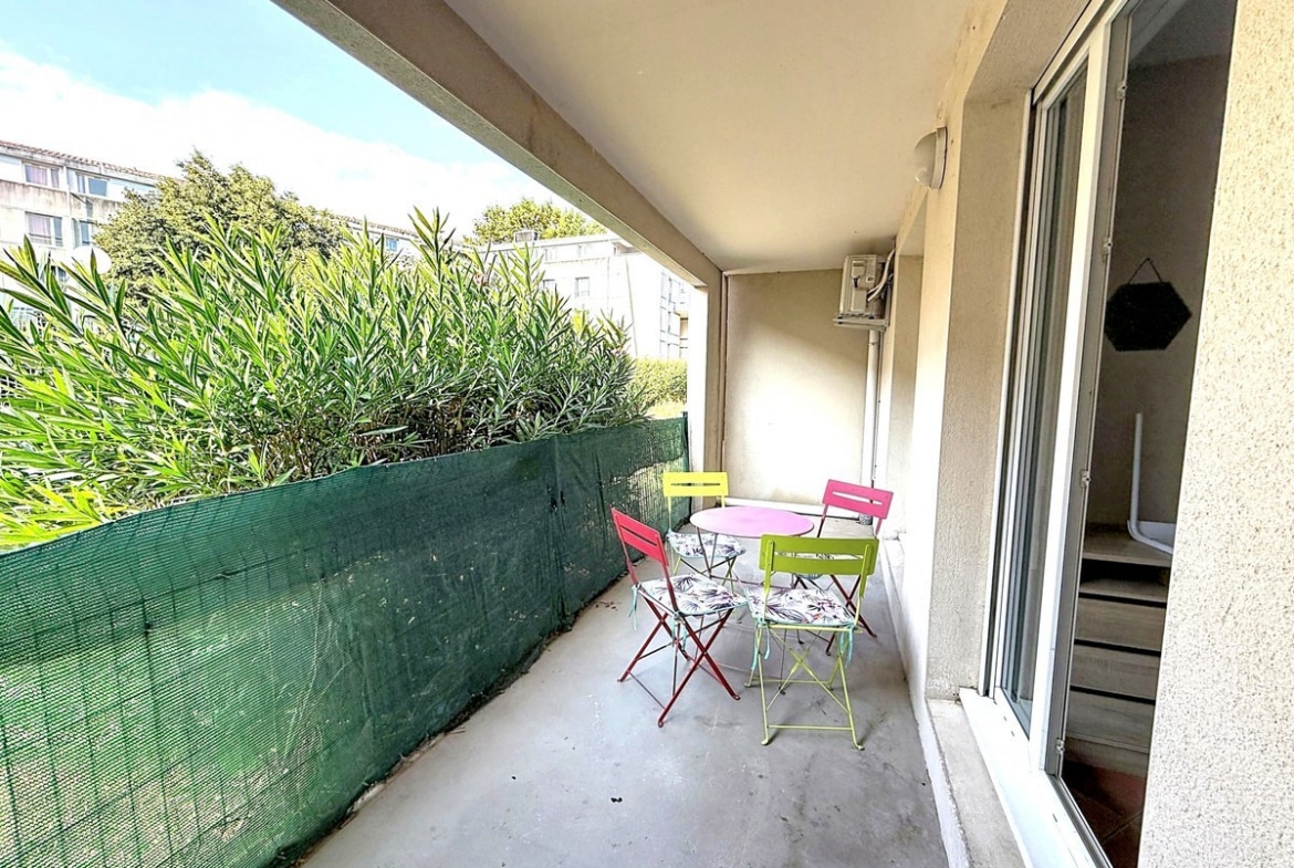 Terrasse couverte de plain-pied avec trois chaises pliantes en métal coloré (rouge, jaune, vert) et une table ronde rose, protégée par un brise-vue en filet vert tendu sur toute la longueur. La terrasse est délimitée par un auvent en béton et donne accès à l'appartement par une porte coulissante vitrée, avec des arbustes à feuilles vertes visibles au-delà de la clôture.