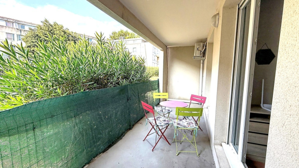 Terrasse couverte de plain-pied avec trois chaises pliantes en métal coloré (rouge, jaune, vert) et une table ronde rose, protégée par un brise-vue en filet vert tendu sur toute la longueur. La terrasse est délimitée par un auvent en béton et donne accès à l'appartement par une porte coulissante vitrée, avec des arbustes à feuilles vertes visibles au-delà de la clôture.