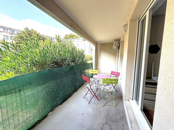 Terrasse couverte de plain-pied avec trois chaises pliantes en métal coloré (rouge, jaune, vert) et une table ronde rose, protégée par un brise-vue en filet vert tendu sur toute la longueur. La terrasse est délimitée par un auvent en béton et donne accès à l'appartement par une porte coulissante vitrée, avec des arbustes à feuilles vertes visibles au-delà de la clôture.