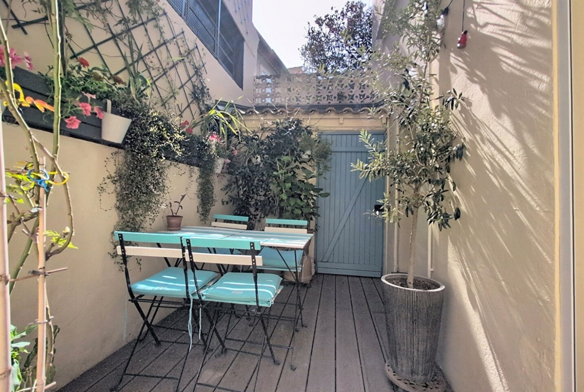 Terrasse extérieure en bois composite avec table et chaises pliantes en métal noir et assises turquoise, entourée de murs enduits beige ornés de plantes grimpantes et de jardinières fleuries. Un olivier en pot cylindrique métallique occupe le premier plan, tandis qu'une porte en bois bleu-vert donne accès à l'impasse au fond de la cour.
