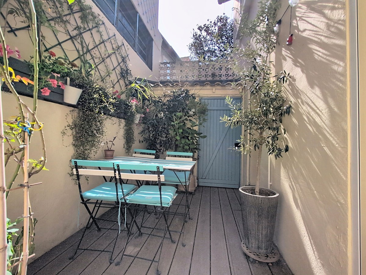 Terrasse extérieure en bois composite avec table et chaises pliantes en métal noir et assises turquoise, entourée de murs enduits beige ornés de plantes grimpantes et de jardinières fleuries. Un olivier en pot cylindrique métallique occupe le premier plan, tandis qu'une porte en bois bleu-vert donne accès à l'impasse au fond de la cour.