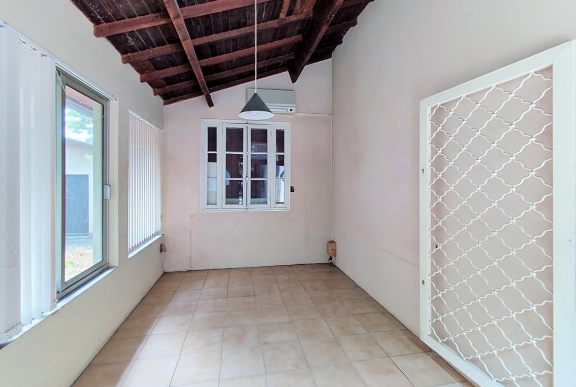 Pièce de type véranda avec charpente en bois brut apparente au plafond, sol carrelé beige et murs blancs, équipée d'un climatiseur mural et d'une suspension noire conique. Une porte-fenêtre métallique blanche avec grille décorative en treillis donne sur l'extérieur, et une grande baie vitrée à jalousies occupe le mur gauche.