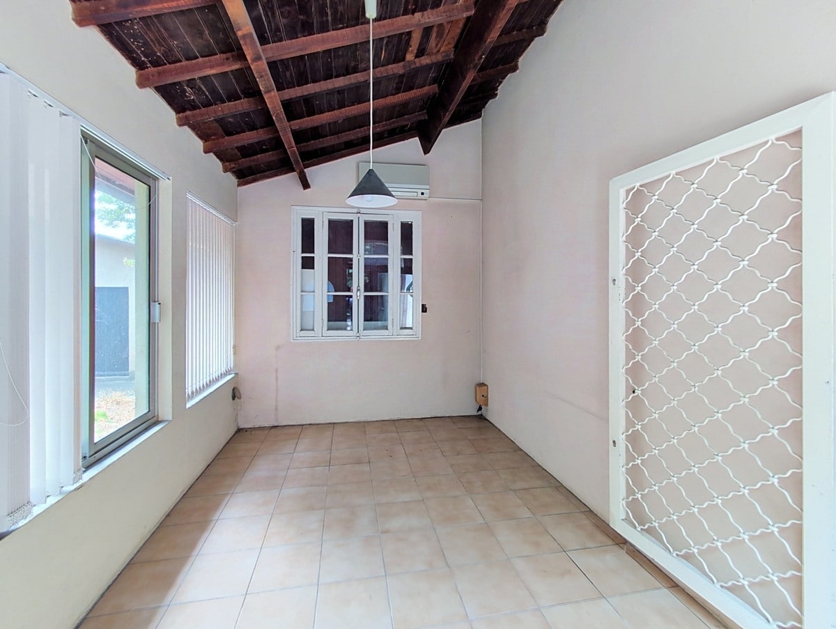 Pièce de type véranda avec charpente en bois brut apparente au plafond, sol carrelé beige et murs blancs, équipée d'un climatiseur mural et d'une suspension noire conique. Une porte-fenêtre métallique blanche avec grille décorative en treillis donne sur l'extérieur, et une grande baie vitrée à jalousies occupe le mur gauche.