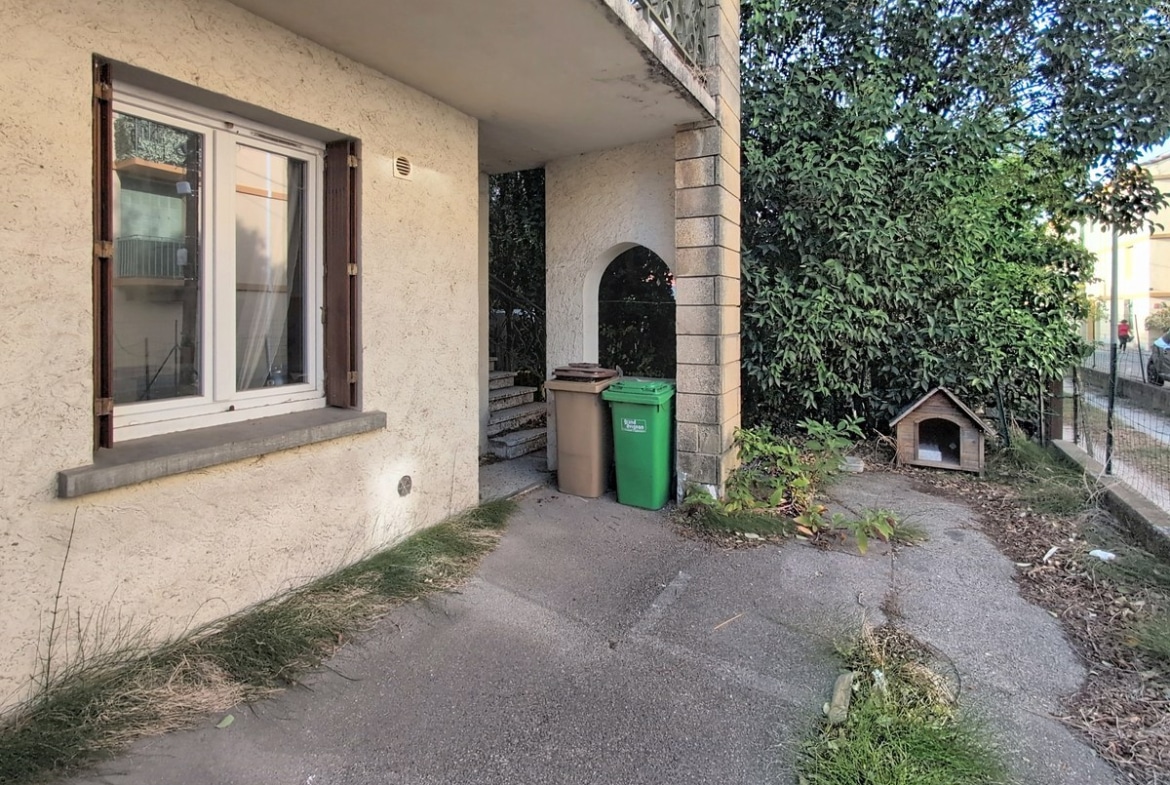 Façade enduite de couleur beige avec fenêtre à volets bois marron, escalier en béton menant à une entrée en arc, deux poubelles de tri sélectif marron et verte placées sous l'escalier. Chemin d'accès en béton longé d'une végétation dense avec un petit abri pour animal de compagnie visible à droite.