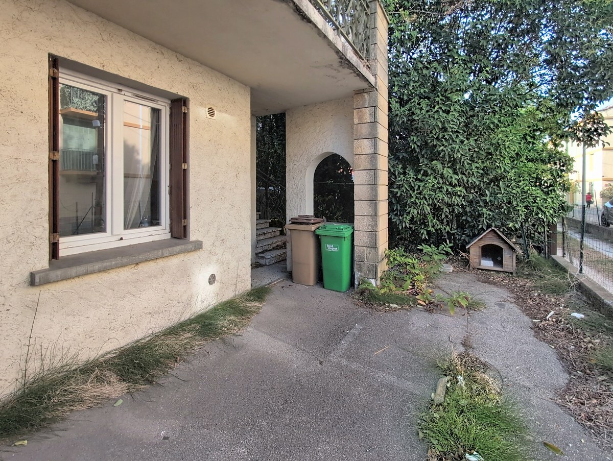 Façade enduite de couleur beige avec fenêtre à volets bois marron, escalier en béton menant à une entrée en arc, deux poubelles de tri sélectif marron et verte placées sous l'escalier. Chemin d'accès en béton longé d'une végétation dense avec un petit abri pour animal de compagnie visible à droite.