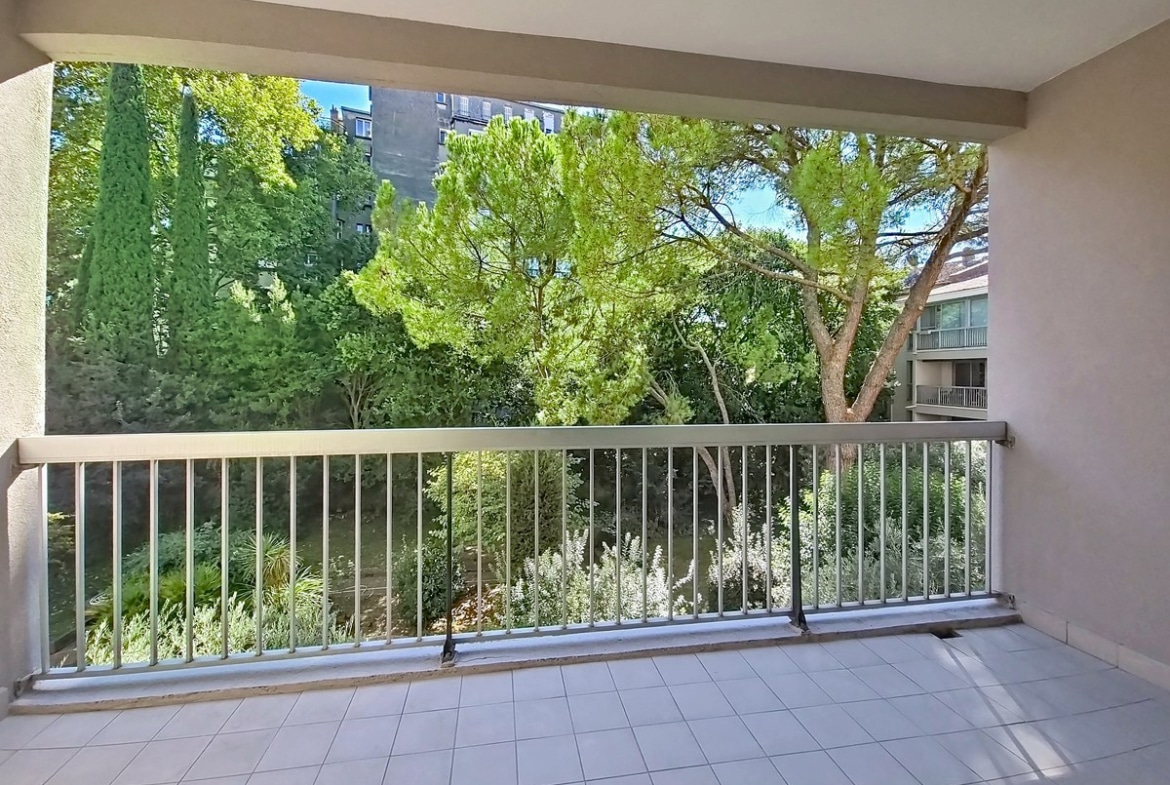 Grand balcon carrelé en blanc avec une garde-corps métallique grise, donnant sur un espace vert planté de pins parasols, de cyprès et d'arbustes méditerranéens. En arrière-plan, un immeuble résidentiel de plusieurs étages avec des balcons est partiellement visible derrière la végétation.