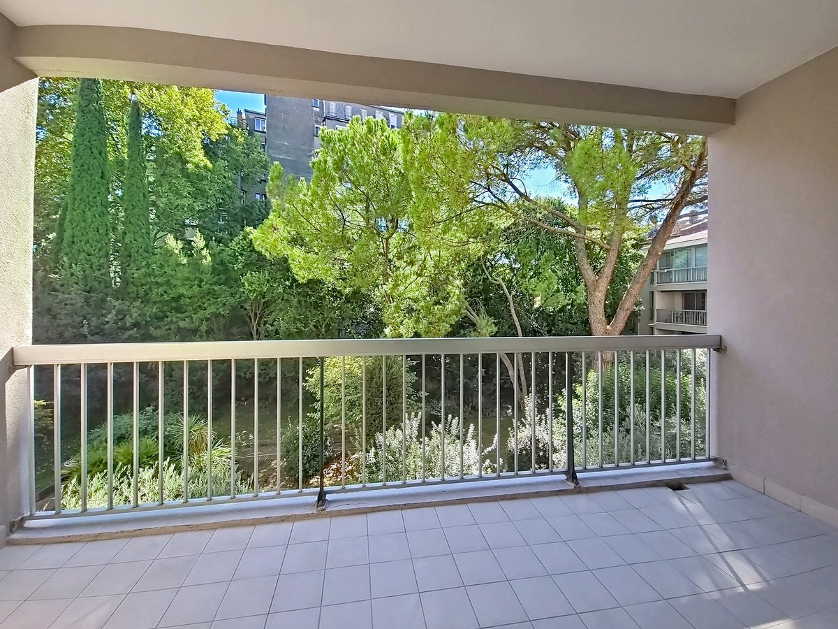Grand balcon carrelé en blanc avec une garde-corps métallique grise, donnant sur un espace vert planté de pins parasols, de cyprès et d'arbustes méditerranéens. En arrière-plan, un immeuble résidentiel de plusieurs étages avec des balcons est partiellement visible derrière la végétation.