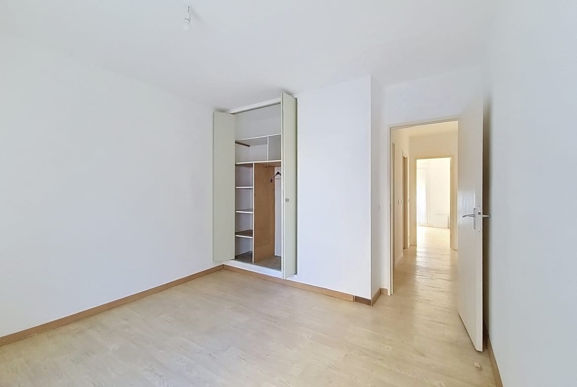 Chambre vide avec un parquet stratifié clair et des murs entièrement blancs, dotée d'un placard encastré à portes coulissantes partiellement ouvert révélant des étagères en bois et une tringle à vêtements. Une porte ouverte en fond de pièce donne sur un couloir lumineux menant vers une autre pièce.