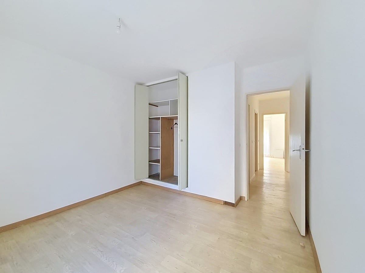 Chambre vide avec un parquet stratifié clair et des murs entièrement blancs, dotée d'un placard encastré à portes coulissantes partiellement ouvert révélant des étagères en bois et une tringle à vêtements. Une porte ouverte en fond de pièce donne sur un couloir lumineux menant vers une autre pièce.