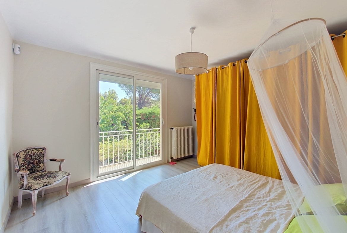 Chambre double avec parquet stratifié clair, équipée d'un lit double sous baldaquin en voile blanc et rideaux jaune moutarde. Une porte-fenêtre coulissante ouvre sur un balcon avec garde-corps en fer forgé blanc, donnant sur une végétation dense de pins et arbres méditerranéens.