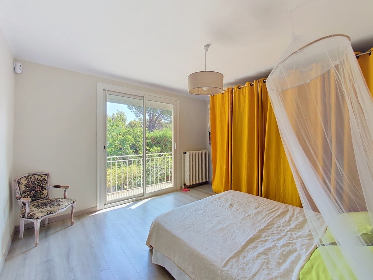 Chambre double avec parquet stratifié clair, équipée d'un lit double sous baldaquin en voile blanc et rideaux jaune moutarde. Une porte-fenêtre coulissante ouvre sur un balcon avec garde-corps en fer forgé blanc, donnant sur une végétation dense de pins et arbres méditerranéens.
