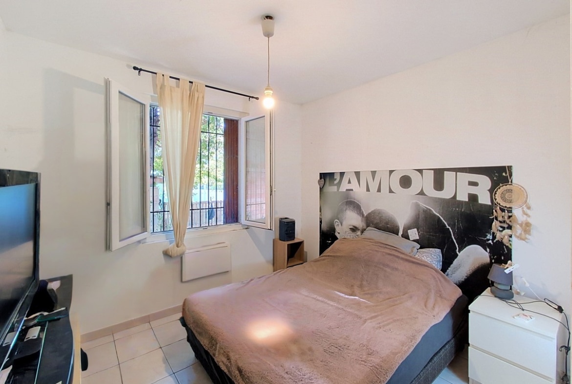 Chambre avec lit double à sommier noir et couverture marron clair, tête de lit constituée d'une affiche noir et blanc portant l'inscription « L'AMOUR ». Fenêtre à deux vantaux avec barreaux et rideau beige, table de chevet blanche IKEA et télévision à écran plat posée à gauche.