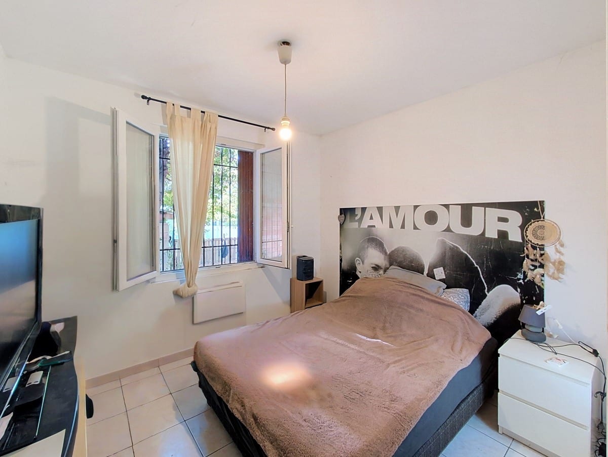 Chambre avec lit double à sommier noir et couverture marron clair, tête de lit constituée d'une affiche noir et blanc portant l'inscription « L'AMOUR ». Fenêtre à deux vantaux avec barreaux et rideau beige, table de chevet blanche IKEA et télévision à écran plat posée à gauche.