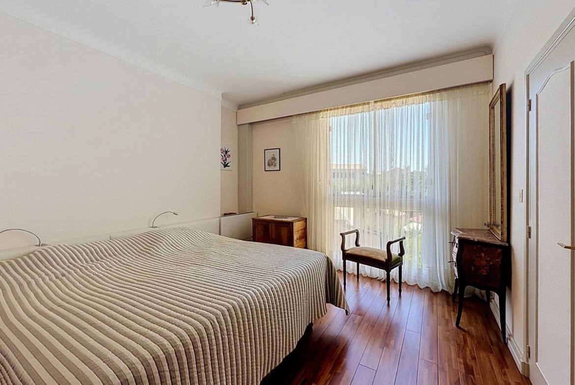 Chambre à coucher avec sol en parquet en bois foncé, meublée d'un lit double avec couvre-lit rayé beige et gris, d'une commode en bois et d'un miroir doré, éclairée par une large baie vitrée à voilages blancs ouvrant sur un balcon. Les murs sont entièrement blancs et un placard encastré est visible sur la droite.