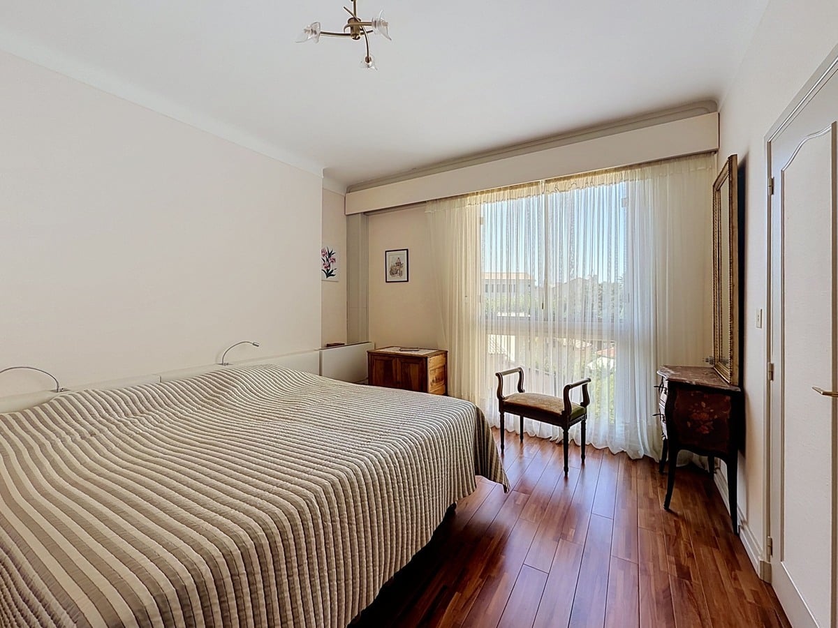 Chambre à coucher avec sol en parquet en bois foncé, meublée d'un lit double avec couvre-lit rayé beige et gris, d'une commode en bois et d'un miroir doré, éclairée par une large baie vitrée à voilages blancs ouvrant sur un balcon. Les murs sont entièrement blancs et un placard encastré est visible sur la droite.