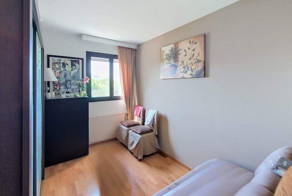 Chambre aux murs beige clair avec parquet en bois clair, équipée d'une grande commode noire placée devant une fenêtre à double vitrage donnant sur la verdure. Un tableau représentant des citrons et un pot en terre orne le mur, et deux fauteuils habillés de tissu clair sont disposés près de la fenêtre.