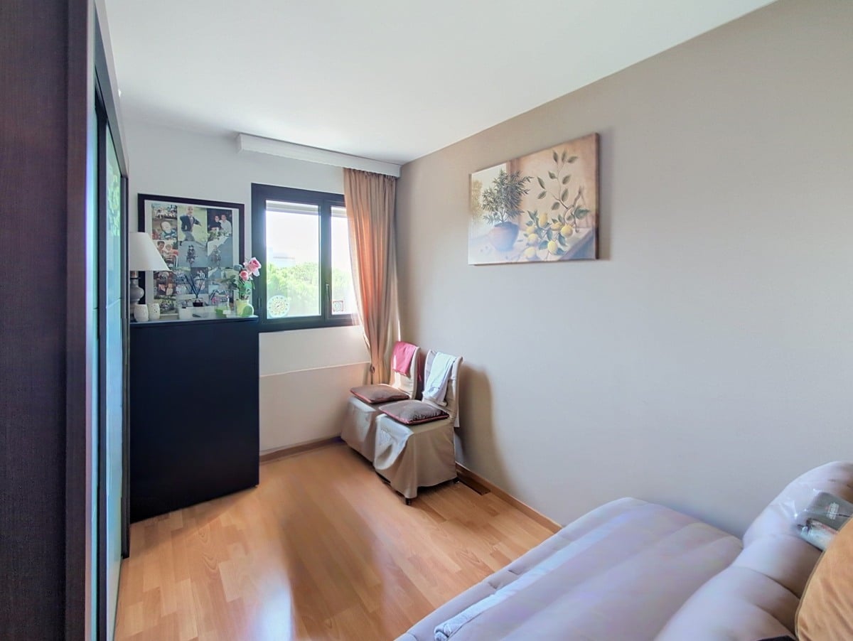 Chambre aux murs beige clair avec parquet en bois clair, équipée d'une grande commode noire placée devant une fenêtre à double vitrage donnant sur la verdure. Un tableau représentant des citrons et un pot en terre orne le mur, et deux fauteuils habillés de tissu clair sont disposés près de la fenêtre.