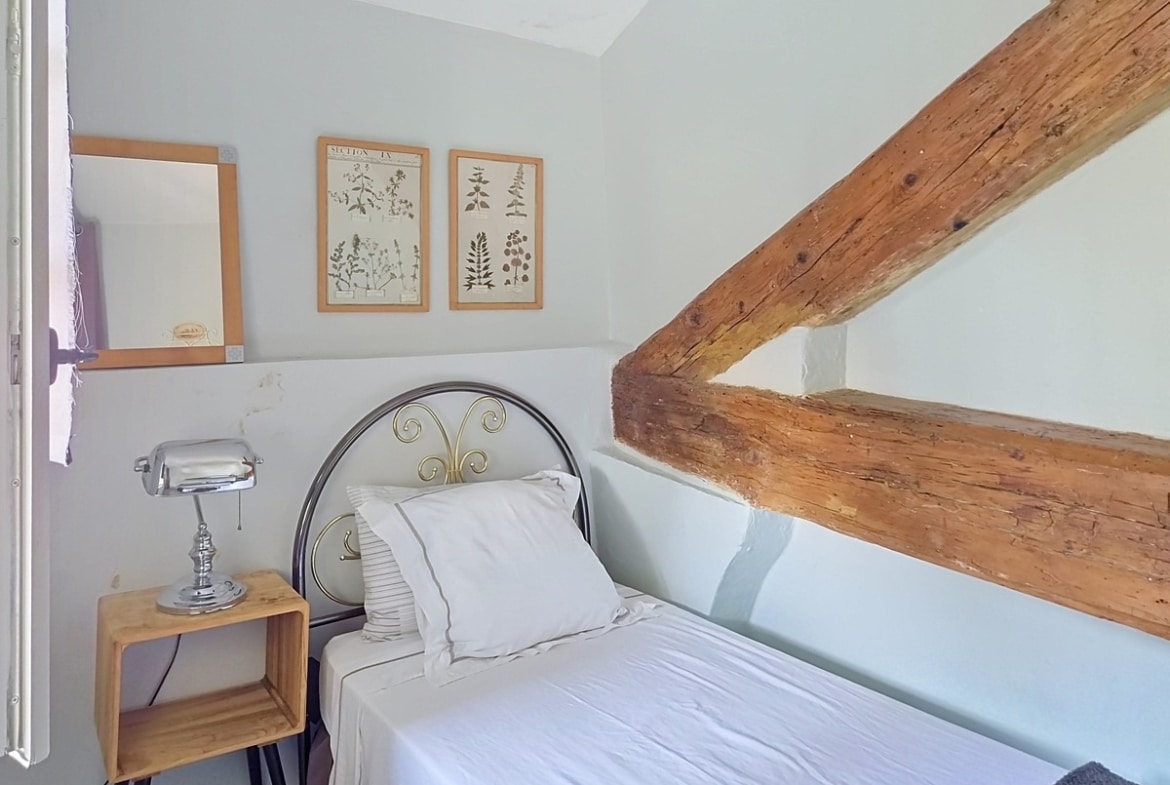 Petite chambre sous combles avec deux poutres en bois brut apparent en diagonale, murs peints en blanc cassé et lit simple à tête de lit en fer forgé avec literie blanche. Table de chevet en caisse de bois brut avec lampe chromée style industriel, deux cadres botaniques encadrés en bois naturel et un petit miroir fixés au mur.