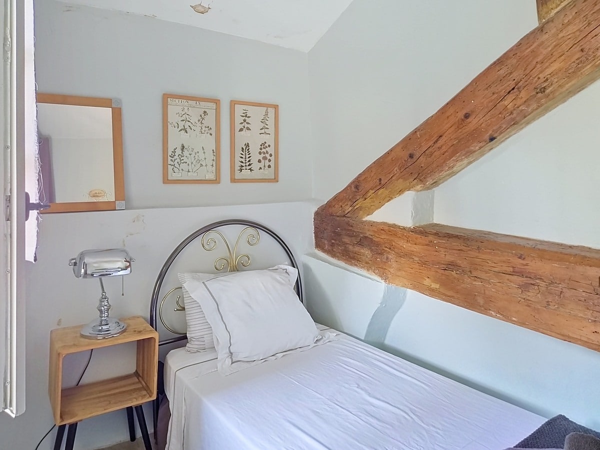 Petite chambre sous combles avec deux poutres en bois brut apparent en diagonale, murs peints en blanc cassé et lit simple à tête de lit en fer forgé avec literie blanche. Table de chevet en caisse de bois brut avec lampe chromée style industriel, deux cadres botaniques encadrés en bois naturel et un petit miroir fixés au mur.