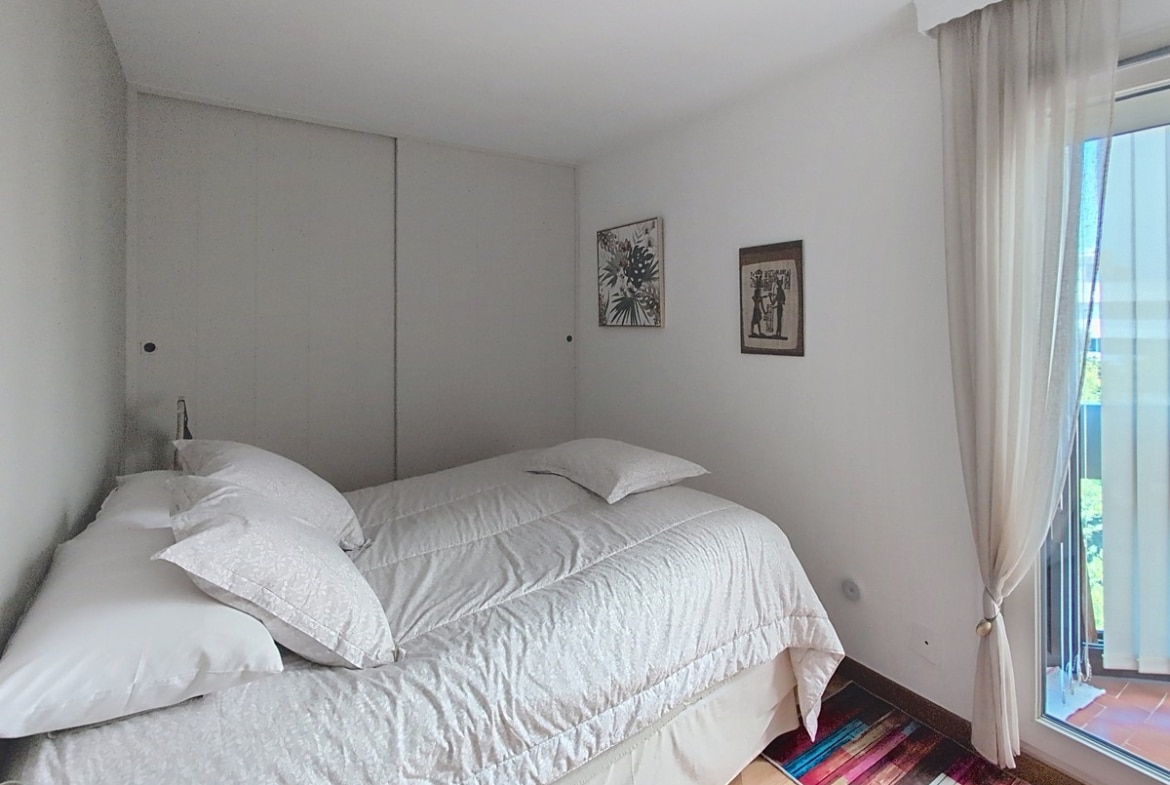 Chambre avec murs blancs et parquet bois clair, dotée d'un grand placard coulissant sur toute la largeur du mur de tête de lit. Un lit double aux draps blancs est positionné face à une baie vitrée habillée de rideaux blancs transparents ouvrant sur une terrasse carrelée en terre cuite.