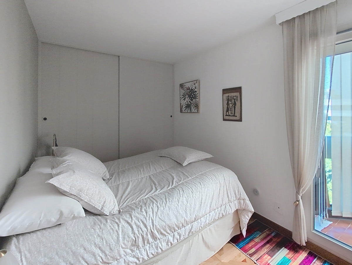Chambre avec murs blancs et parquet bois clair, dotée d'un grand placard coulissant sur toute la largeur du mur de tête de lit. Un lit double aux draps blancs est positionné face à une baie vitrée habillée de rideaux blancs transparents ouvrant sur une terrasse carrelée en terre cuite.