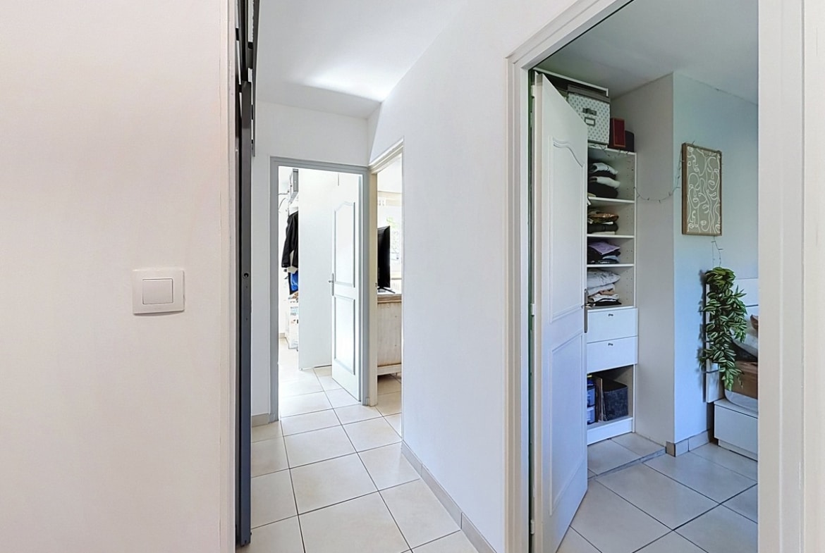 Couloir blanc avec carrelage gris clair donnant sur plusieurs pièces, dont une chambre avec placard ouvert à étagères blanches contenant du linge plié. Porte grise entrouverte en premier plan et ouverture vers une pièce lumineuse en fond de couloir.