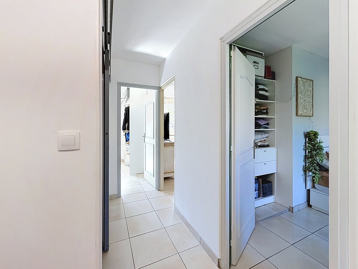 Couloir blanc avec carrelage gris clair donnant sur plusieurs pièces, dont une chambre avec placard ouvert à étagères blanches contenant du linge plié. Porte grise entrouverte en premier plan et ouverture vers une pièce lumineuse en fond de couloir.