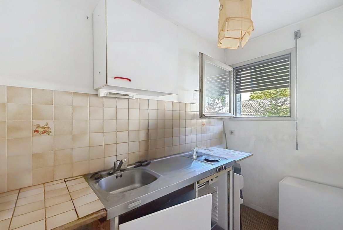 Cuisine étroite avec carrelage mural beige, évier en inox double bac et plaque de cuisson intégrée sur plan de travail carrelé, associés à un réfrigérateur blanc visible sur le côté. Une fenêtre à jalousies métalliques laisse entrer la lumière naturelle sur l'espace cuisine, au-dessus duquel est fixé un meuble haut blanc.